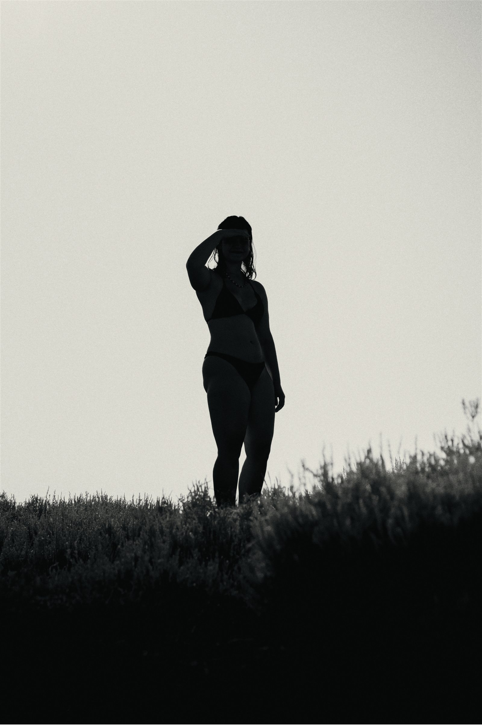 A woman in swimwear standing in tall grass, silhouetted against a bright sky