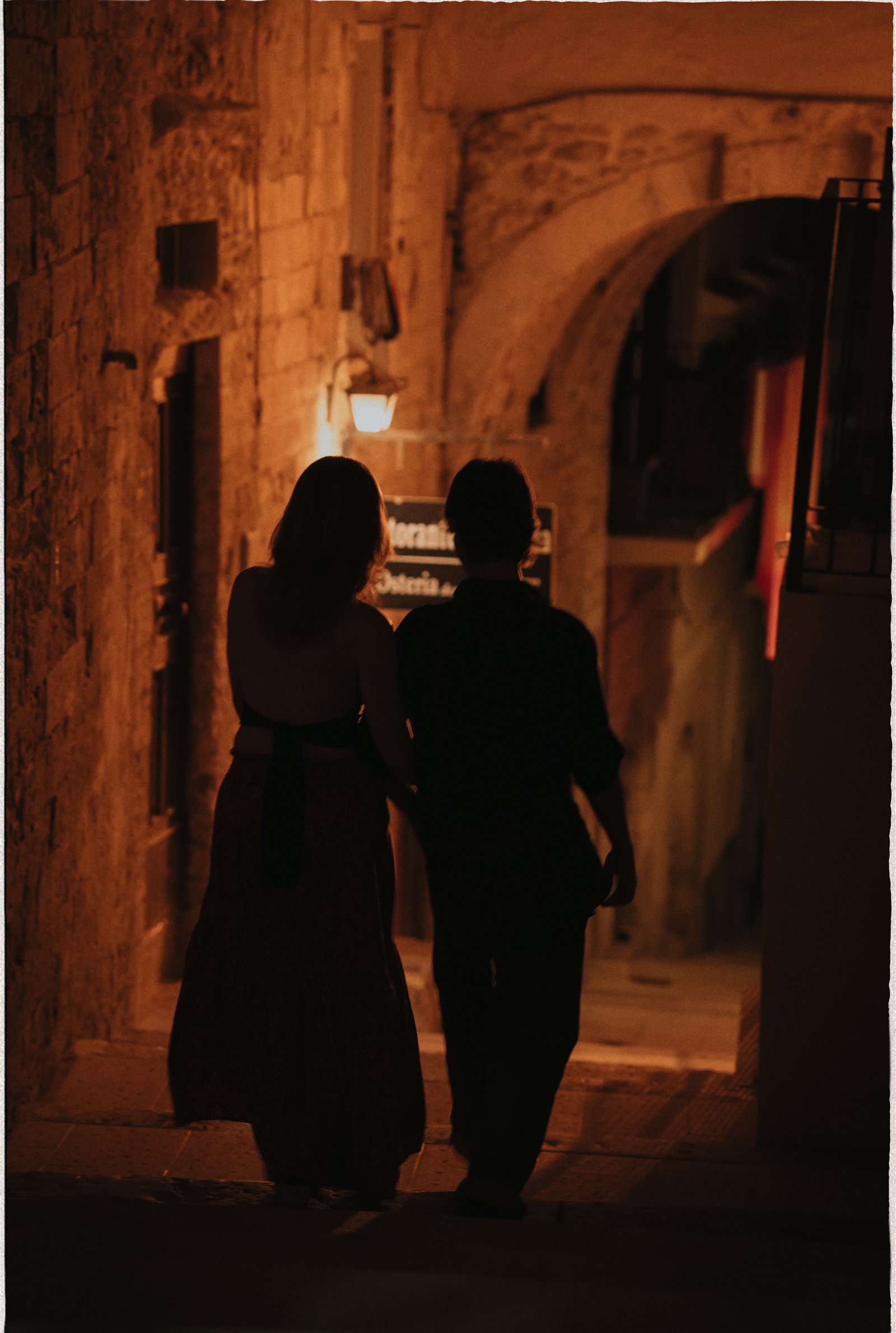 A couple silhouetted in a warm-lit stone alley at night